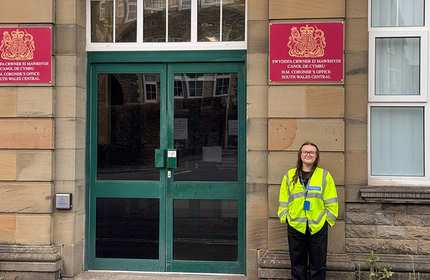 A young woman in glasses wearing a high visibility jacket stands next to the entrance to the HM Coroner's Office for South Wales Central