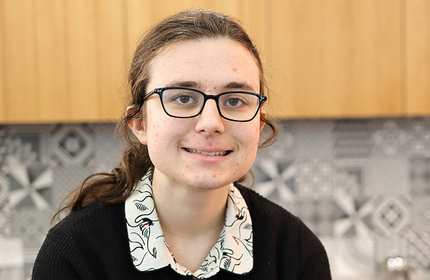 A young woman, wearing glasses and a floral blouse, sits in front of a textured wooden background