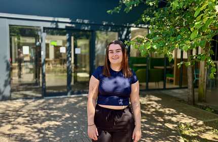 A young woman in a dark top and bottoms stands in front of a shaded building entrance