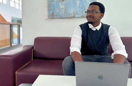 A young person in a shirt and vest works on a laptop while sat in a maroon leather sofa
