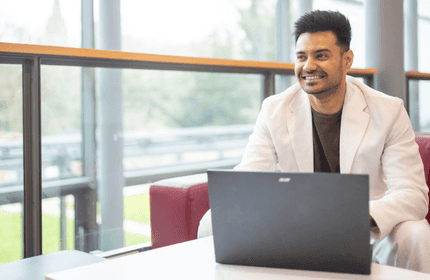 A young man in a white suit sits at a table looking up from their laptop