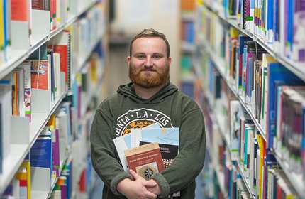 Sammy Chapman stands in between library bookshelves holding a selection of books