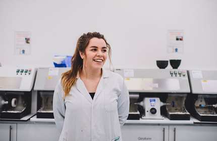 A woman in a white lab coat smiles while standing in front of laboratory equipment. The room is bright and the equipment is lined up along a countertop.