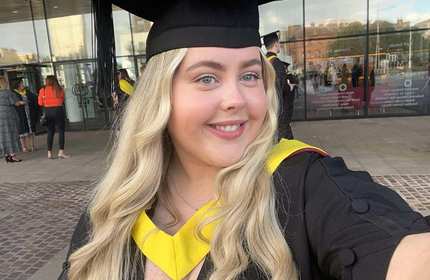 A person wearing graduation robes and cap. In the background are other people wearing similar robes.