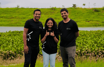 Three people in black shirts smile outdoors by a lake and greenery. The person in the middle gives a thumbs-up. Its a bright, slightly cloudy day, with a grassy hill in the background.