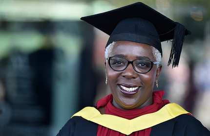 A smiling person wearing a graduation cap and gown with a yellow sash stands outdoors. They have short gray hair and are wearing glasses, with trees blurred in the background.