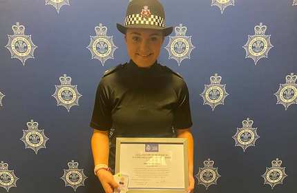 A police officer in uniform holds a certificate and smiles at the camera. The background features a patterned wall with police insignia.