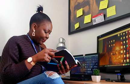 A woman sits at a desk reading a book, surrounded by two computers. Sticky notes are on the wall behind her, and a small plant decorates the desk. She wears a watch and earrings, with her hair in a bun.