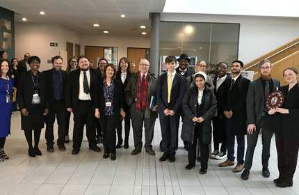 A diverse group of adults posing indoors, some dressed formally. One person holds a shield-shaped award plaque. They stand in a bright hallway with glass doors and stairs in the background.