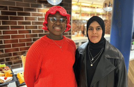 Two people stand together indoors. One wears a red hat and sweater, and glasses. The other wears a black hijab and leather jacket. Behind them is a table with food and a brick wall with shelves.