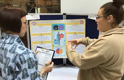 Two women stand in front of a colorful research poster. One woman gestures while explaining, and the other holds a clipboard. The poster contains charts and diagrams related to health and medicine.