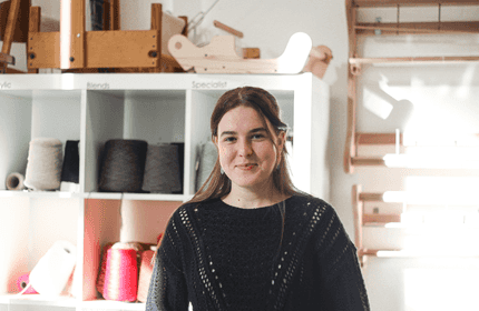 A person with long hair wearing a black sweater stands smiling in front of shelves holding various spools of thread and wooden looms. The room is brightly lit, highlighting the creative workspace.