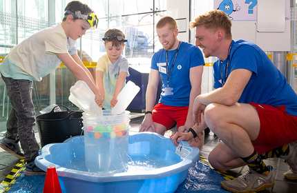 Two children in swimwear and goggles pour water into a bucket in a small pool, assisted by two adults in uniforms. The indoor setting is bright with natural light from large windows. Colorful balls float in the pool.