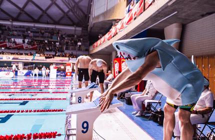 A swimmer in a shark costume leans over a starting block, preparing to dive into an indoor pool during a swim competition. The pool is surrounded by spectators and other competitors.