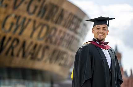 A person wearing a graduation cap and gown. Behind them is the Wales Millennium Centre.