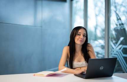 A person sits at a table. In front of them is a laptop computer and an open notepad.
