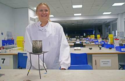 A person smiling in a science lab, wearing a white lab coat. In front of them is a tripod with a metal container. The background has blue chairs and various lab equipment on benches.