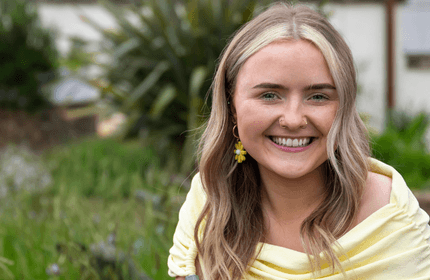 A woman with long blonde hair smiles brightly while sitting outdoors. She is wearing a yellow top and yellow flower earrings. The background features greenery, with blurred plants and a building visible.