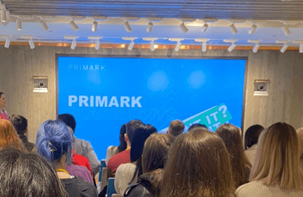 A group of seated people facing a large screen in a presentation room. The screen displays the Primark logo and the text PLACES IT? in a brightly lit environment. The audience is diverse, with varying hair colors and styles.