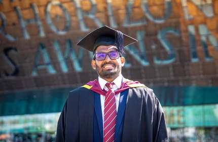A man wearing a graduation cap and gown stands smiling. In the background is the Wales Millennium Centre.
