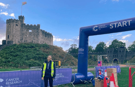 A person in a high-visibility vest stands near a starting line arch for a Cancer Research UK event. The scene is set near a historic stone castle with a blue sky backdrop.