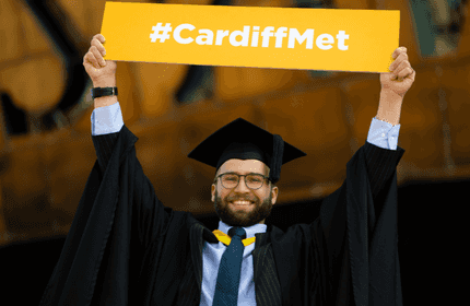 A graduate wearing a cap and gown joyfully holds up a yellow sign with #CardiffMet written on it.
