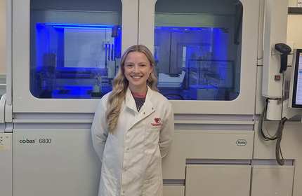 A woman in a lab coat stands smiling in front of a large scientific machine with blue lighting. She has long blonde hair and is in a laboratory setting.