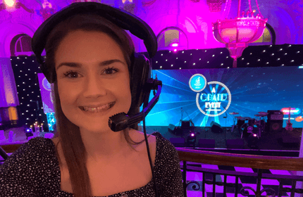 A woman wearing a headset smiles at the camera in a venue with purple lighting. Behind her is a stage with a large screen displaying the text Grand Event. Musical instruments are visible on stage.