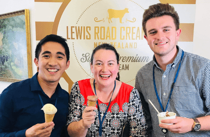 Three people smiling and holding ice cream cones stand in front of a Lewis Road Creamery sign. The person in the center wears a red-patterned shirt, while the others are in dark shirts. A painting of cows is visible on the left.