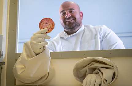 A scientist in a white lab coat and protective gloves holds up a petri dish with colonies inside a laboratory containment unit. He is smiling and looks focused on his work.