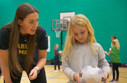 A young woman with long hair is smiling and talking to a blonde girl who is holding a basketball. They are in a gymnasium with other children playing in the background.