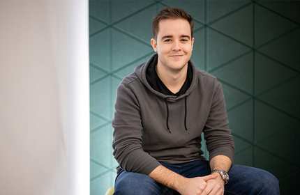 A person wearing a gray hoodie and blue jeans is sitting in front of a geometric-patterned wall. The individual is smiling with hands clasped and is positioned slightly to the right against the textured backdrop.
