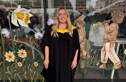 A young woman wearing a graduation gown stands smiling. In the background are glass windows decorated with illustrations of flowers.