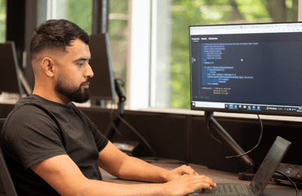 A man with short dark hair and a beard is sitting at a desk in an office, typing on a laptop. A large monitor in front of him displays code. The room has several desks with computers and large windows showing greenery outside.