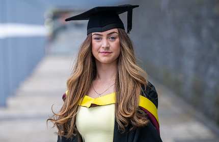 A young woman wearing a graduation gown and cap stands outdoors on a pathway. She has long, wavy hair and is dressed in a yellow top under the gown. The background is slightly blurred, with a stone wall visible.
