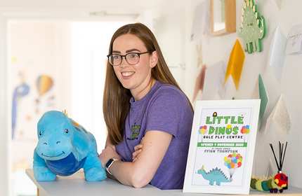 A smiling person with glasses leans on a counter next to a blue dinosaur plush toy. A framed poster announces the opening of Little Dinos Role Play Centre on December 6, 2020. The background displays wall decorations and papers.