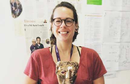 A person wearing glasses and a red shirt smiles while holding a gold award mask. They are standing in front of a wall covered with papers and clippings.
