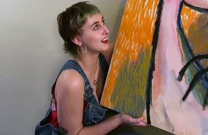 A person with short hair and hoop earrings, wearing overalls and a floral top, crouches while holding a colorful abstract painting. They look upwards with a smile, set against a neutral background.