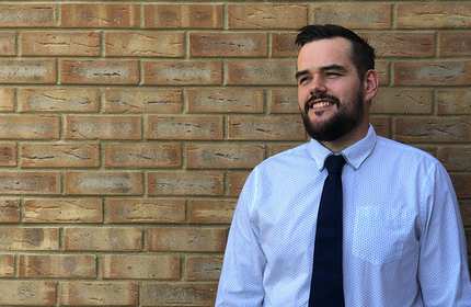 A person wearing a shirt and tie stands against the background of a brick wall.