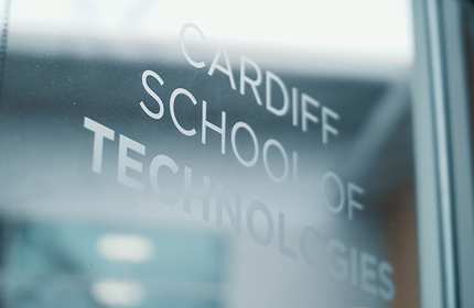 A glass door with the words Cardiff School of Technologies etched on it, viewed from an angle. The background appears blurred, hinting at an indoor space on the other side of the door.