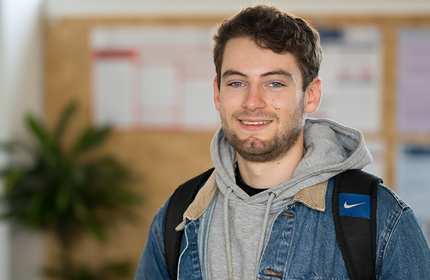 A man with short hair, wearing a denim jacket over a gray hoodie and carrying a backpack, smiles at the camera. The background is blurred, showing some bulletin boards and a leafy plant.