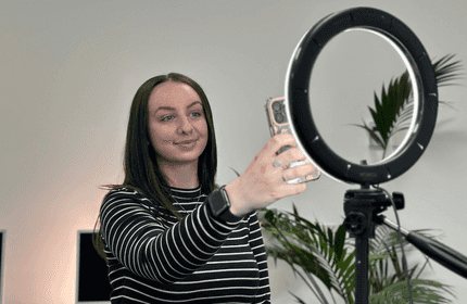 A person with long hair and a striped shirt uses a smartphone, with a ring light on a stand in front of them. There are plants and computer monitors in the background.