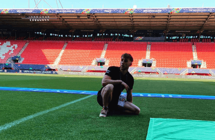 A person kneels on a sports field in an empty stadium with red seats. The stadium is decorated with UEFA Nations League banners. The blue and green banners on the ground indicate an event or ceremony setup.