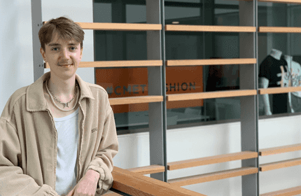 A person with short brown hair and a light jacket stands indoors by a wooden railing. Behind them is a glass wall with a sign and a mannequin displaying a graphic T-shirt.