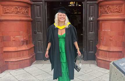 A woman wearing a green dress and graduation gown with a cap stands smiling in front of a red brick building. She holds a patterned bag and appears to be celebrating her graduation.