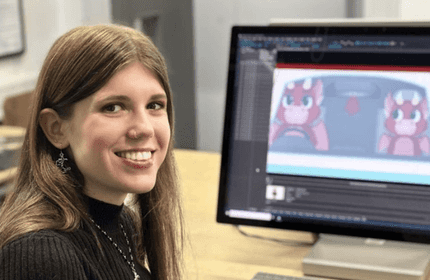 A person with long hair smiles at the camera while sitting at a desk. In the background, a computer screen displays an animation of two cartoon characters driving a car.