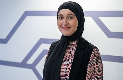 A person wearing a black hijab and a plaid shirt smiles softly while standing in front of a geometric wall design.