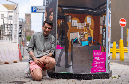 A person in a gray shirt and pink shorts kneels next to a cylindrical public display with various posters and images. The street setting includes a P parking sign and yellow bollards.
