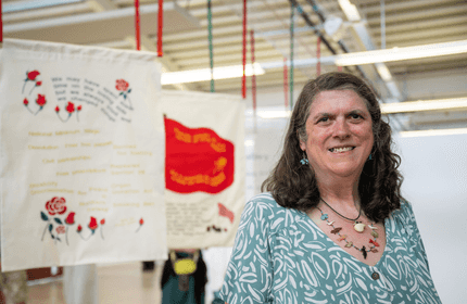 A woman in a patterned green shirt is smiling at the camera. In the background, there are hanging fabric banners with various designs and text. The setting appears to be an art exhibition space with overhead lighting.