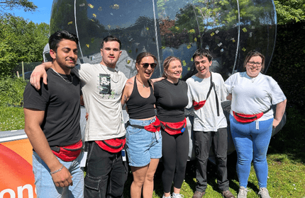 A group of six people smiling and standing close together outdoors in daylight. Some wear fanny packs, and theres a transparent, inflated structure in the background with trees and clear sky visible.